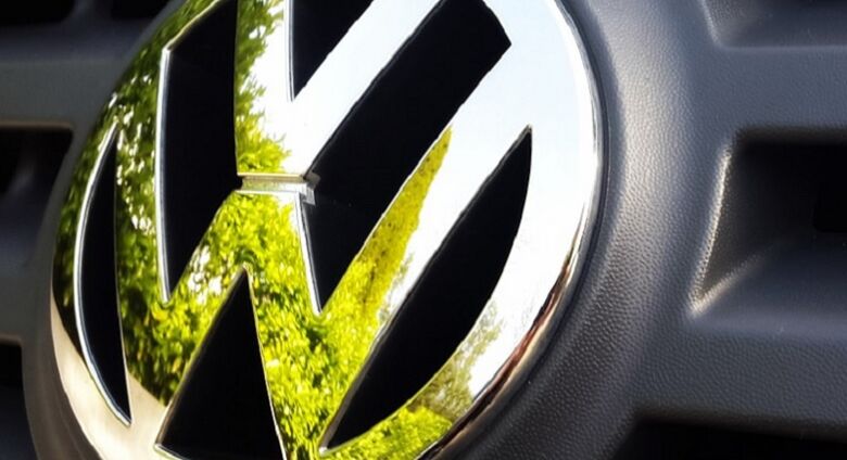 Nahaufnahme eines VW Logos auf einem Auto. Durch die Spiegelung erkennt man Bäume. Stellt die Verbindung zwischen Mobilität, Umwelt und Nachhaltigkeit dar.